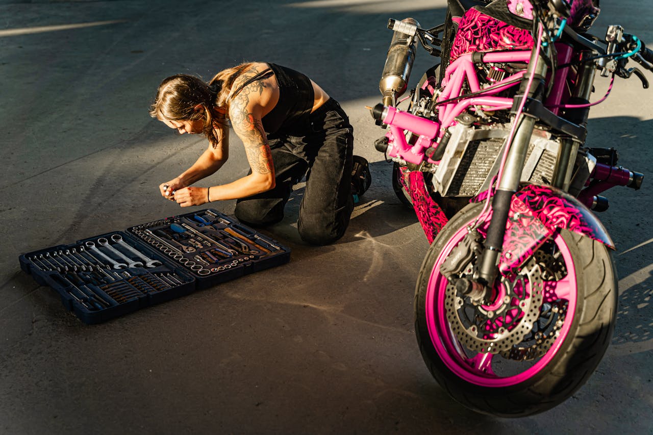 gallery-2 A woman with tattoos kneeling on a concrete floor, working on a pink motorcycle using a toolbox.